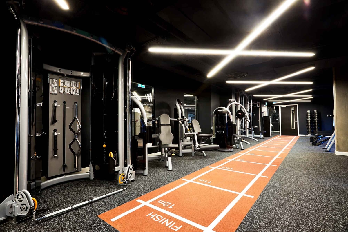 Modern gym with exercise machines and weights along the walls. A bright, orange-tiled running track with distance markers runs through the center. Overhead, contemporary lighting fixtures illuminate the dark-themed interior.